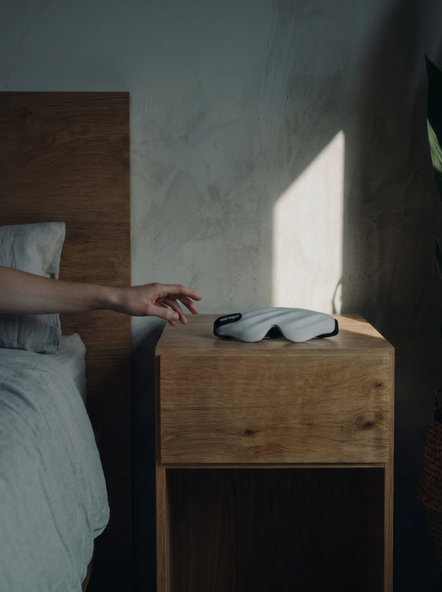 Person reaching for a white eye mask on a wooden nightstand in a bedroom.